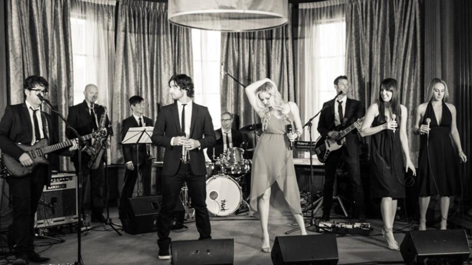 A group of people is performing on stage, showcasing a musical performance. They are dressed in various clothing styles, including suits, while playing instruments such as a guitar and drums. The setting appears to be an indoor concert with a curtain backdrop. The image has a black and white aesthetic, adding to its artistic flair.