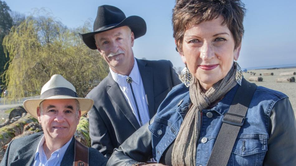The image is a medium shot of three people posing outdoors on a beach on a sunny day. In the foreground, on the left, is a man wearing a light-colored fedora with a dark band, a grey blazer over a light blue collared shirt, and a brown guitar strap across his chest. He is looking directly at the camera with a slight smile and has short, dark hair.