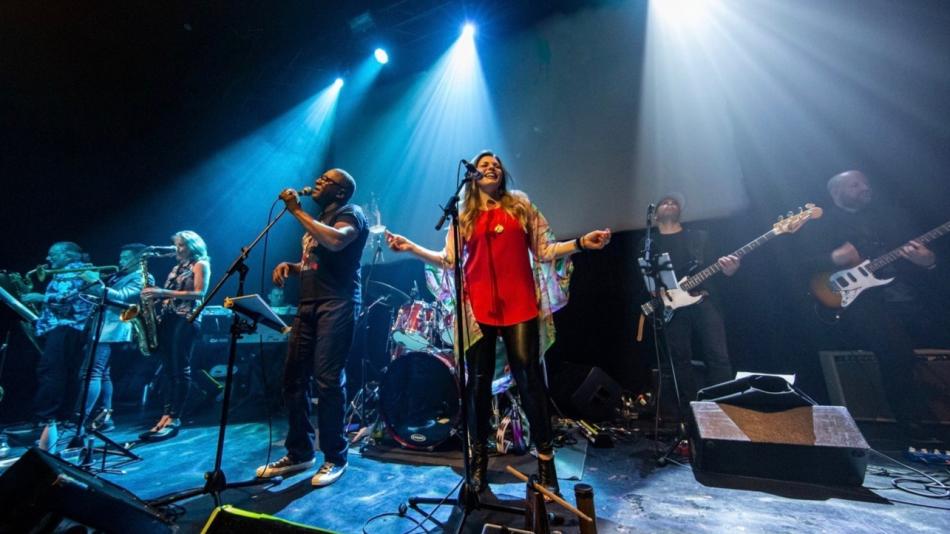 The image captures a live musical performance on a dimly lit stage. The primary focus is on a band with several musicians, illuminated by bright spotlights from above. The stage floor appears to be a dark, textured material, possibly wood or concrete, with some cables and equipment visible.