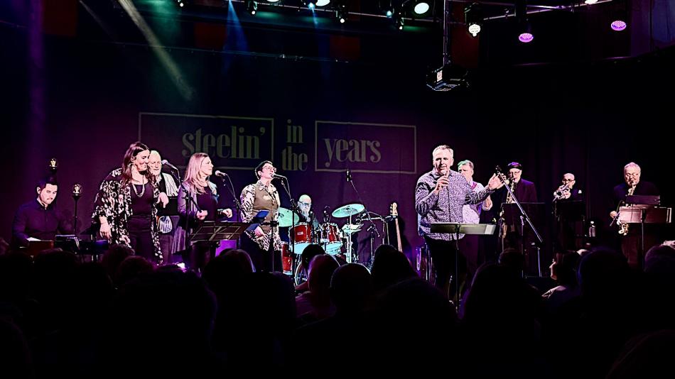 The image captures a live musical performance taking place on a stage. The background features a dark curtain with a large, illuminated banner that reads "steelin' in the years" in a stylized, outlined font. The banner has a purple hue, contrasting with the stage lighting.