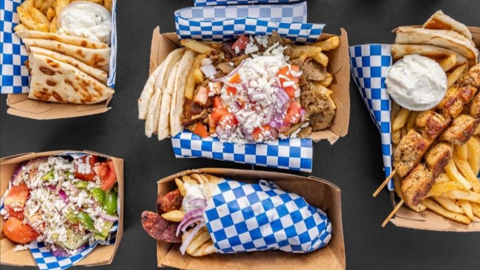 The image displays a spread of five distinct takeout containers filled with various Greek food items, arranged on a dark, textured surface. The containers are primarily made of light brown cardboard, with a blue and white checkered paper lining peeking out from the edges of most of them.