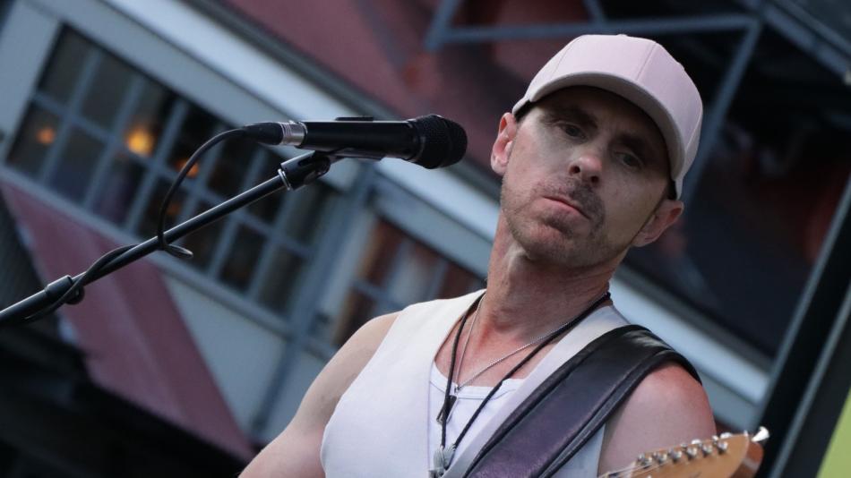 A male musician is performing on stage, holding an electric guitar and singing into a microphone. He is wearing a white tank top, a light pink baseball cap, and two silver necklaces with pendants. The guitar is a light brown color with a dark fretboard. The microphone is positioned in front of his face. The background is blurred but appears to be an outdoor venue with some structures and lights. The overall lighting suggests it is either daytime or early evening. The composition is a medium shot focusing on