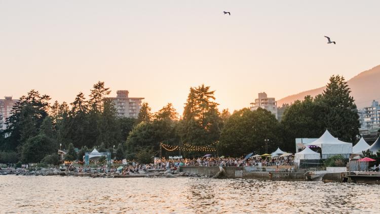 A large crowd gathers on the waterfront, enjoying a summer evening under the setting sun.  The scene is framed by a row of apartment buildings and lush green trees.  The sun casts a warm glow over the water, creating a sense of peace and tranquility.  A boardwalk extends into the water, and several white tents are set up on the shore, suggesting a festival or event.  The scene is alive with laughter and conversation, creating a vibrant and festive atmosphere. 