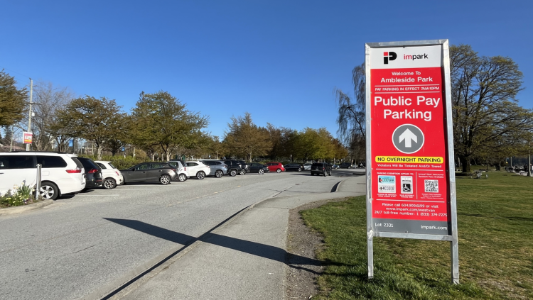 A red parking sign reads "Welcome to Ambleside Park - Public Pay Parking" with a parking lot and blue sky in the background.
