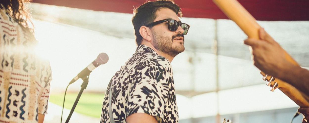 The main subject is a man in the mid-ground, seen from the chest up and in profile, looking to his right. He has dark hair, a beard, and is wearing dark-rimmed sunglasses. His shirt is the most striking detail of his attire, featuring a bold black and white pattern that resembles abstract animal prints or ink blots. He is holding a guitar, visible from the shoulder down, and a strap runs diagonally across his chest.