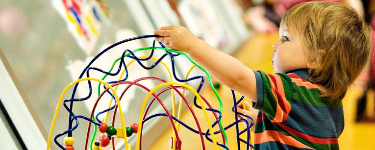 A toddler plays with a toy that has wooden spheres on a maze of brightly coloured wires. 