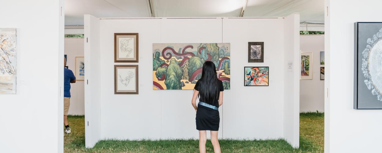 A woman observes artwork displayed in an outdoor exhibition, surrounded by greenery and natural light.