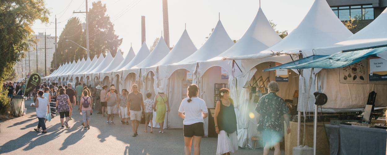 A bustling art market scene with a large group of people strolling along a street filled with colorful tents.