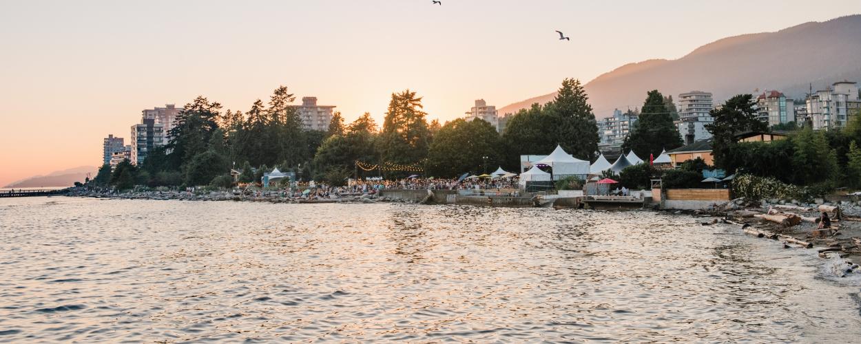 The image captures a scenic view of an event taking place along a shoreline, possibly a festival or outdoor gathering, with a body of water in the foreground. In the background, there are buildings, trees, and what appears to be a mountain range partially obscured by the hazy sunlight. The sky is a soft gradient of colors, suggesting either sunrise or sunset, and two birds are seen flying overhead. 
