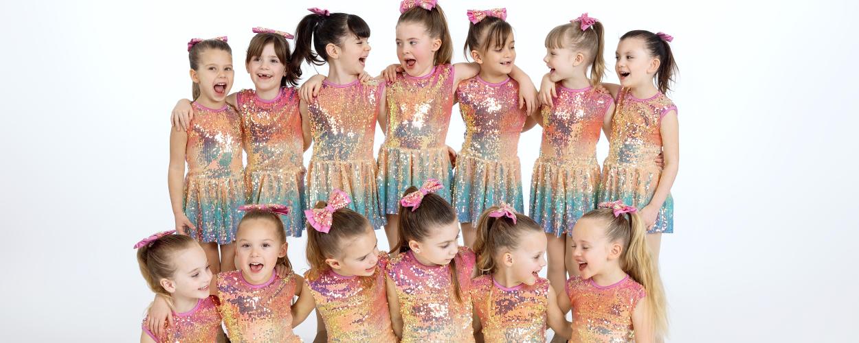 A group of young girls wearing colorful sequinned dresses, smiling and posing together for a photo.