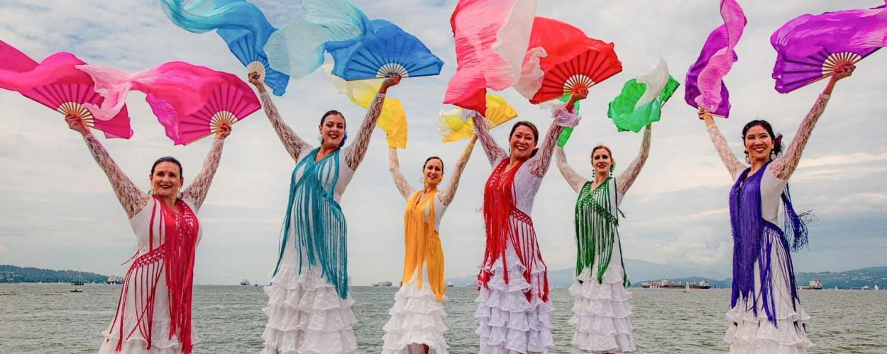 A group of dancers in colorful dresses and fans stands on rocks with the ocean in the background.