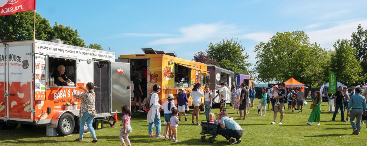A group of people is gathered around a food truck, which is parked in an outdoor setting. The surroundings include grass and trees, with a clear sky overhead. The scene depicts a lively public space where individuals are likely enjoying the food being served from the mobile vendor.