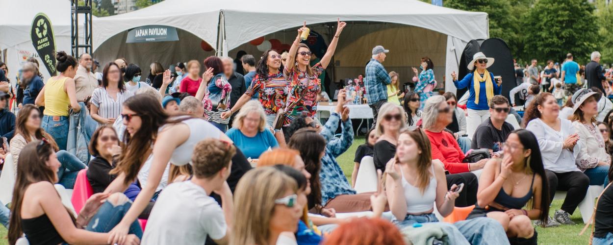 The image depicts a large crowd of people gathered in a field. They are sitting on the grass and appear to be enjoying an outdoor event or festival. The scene includes various individuals, including men, women, and children, dressed in casual clothing and footwear. Tents can be seen set up around the area, contributing to the festive atmosphere.
