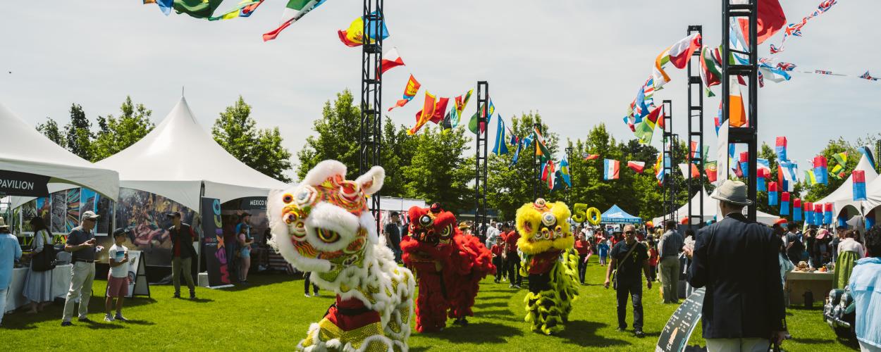 A group of people are gathered at an outdoor event. The setting features a clear sky and lush grass, with trees in the background. Attendees are dressed in various clothing styles and seem to be enjoying activities such as dancing, contributing to the festive atmosphere of the occasion.