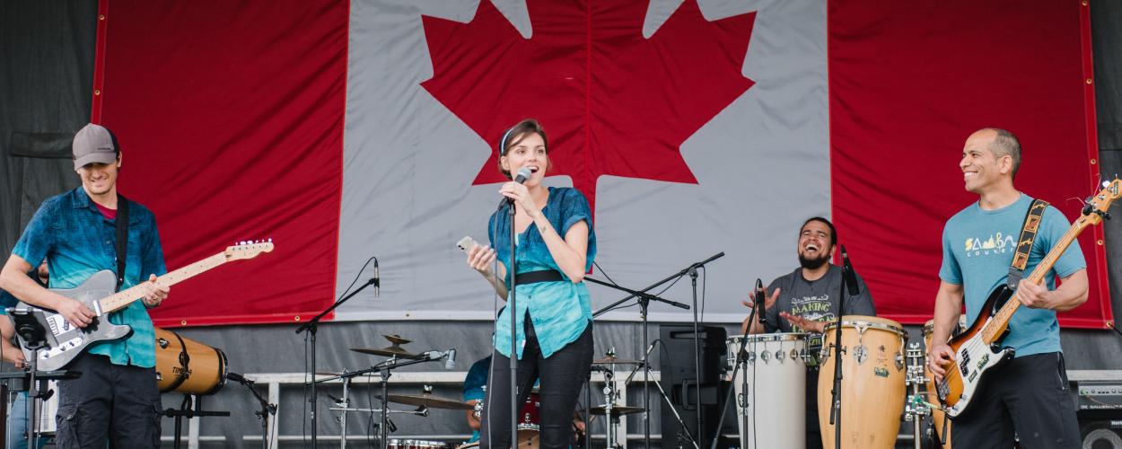 A diverse group of musicians performs on stage, featuring a female singer, male guitarist, bass player, and drummer, with a Canadian flag.