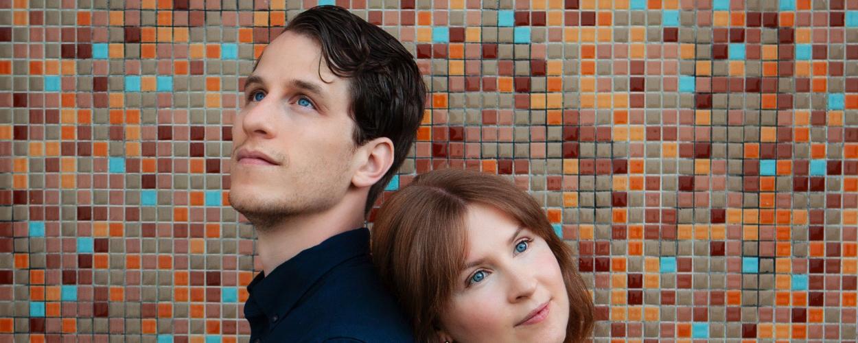 The image is a medium shot portrait of a man and a woman positioned back-to-back against a colorful, mosaic tiled wall. The man is on the left, facing upwards and slightly to the left, with his head tilted back. He has short, dark brown hair, fair skin, and striking blue eyes. He is wearing a dark navy blue collared shirt.