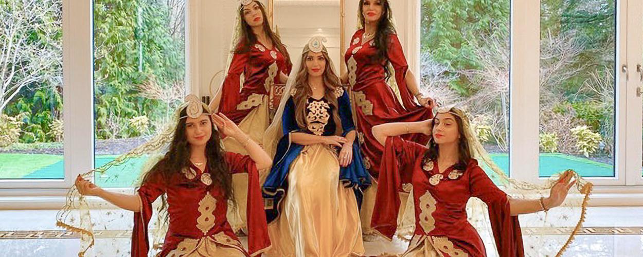 The four surrounding women are dressed in matching elaborate costumes, primarily in rich, deep red velvet with gold accents. Their gowns feature wide, flowing sleeves and ornate gold embroidery on the bodice and waist. Each woman wears a similar headpiece and a sheer, golden veil that flows gracefully around them. Their poses are dynamic and stylized: the two on the left and right are seated with one hand raised near their head, holding the veil, while the other hand is extended outward. 