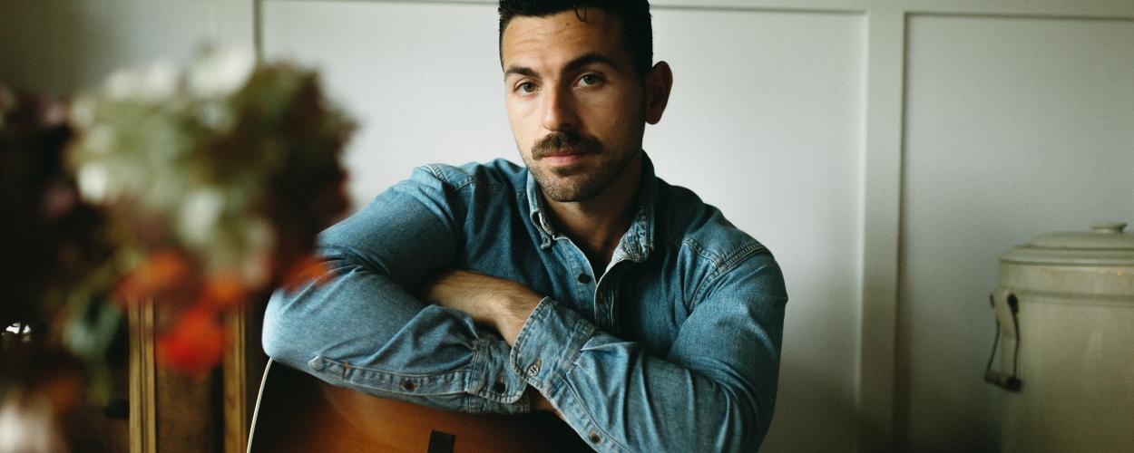 A man with dark, curly hair and a mustache is seated, holding an acoustic guitar. He is wearing a denim shirt, and his arms are crossed over the guitar. The man is looking directly at the camera with a serious expression. The background is a light-colored wall with some molding, and to the right, there is a large, cream-colored ceramic pot with a lid and a handle. In the foreground to the left, a vase of flowers is visible, though it is blurred and out of focus. The lighting is soft and warm, casting a gent