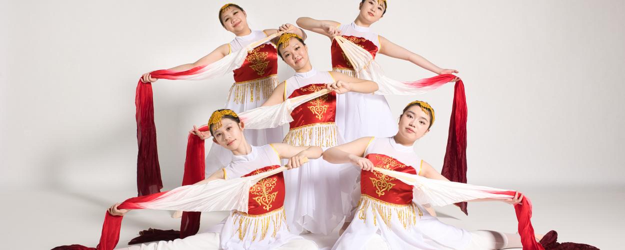 Five young women are positioned in a visually dynamic arrangement against a plain white background, suggesting a studio setting for a performance or photoshoot. They are all dressed in elaborate, traditional-inspired costumes featuring a white base with bright red bodices adorned with intricate gold embroidery and delicate gold fringe.