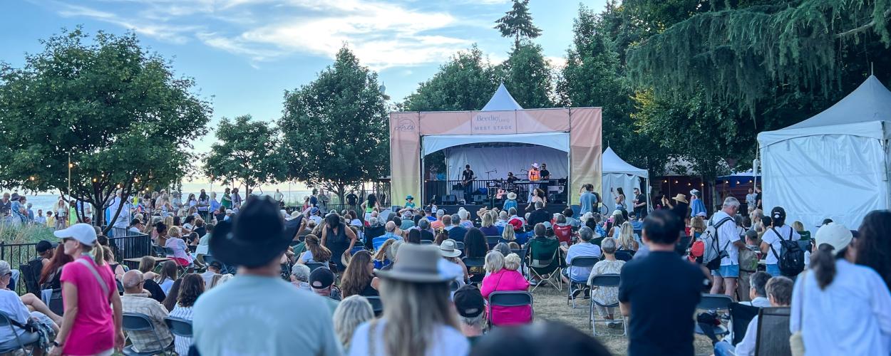 This is a wide-angle shot of an outdoor concert or festival on a clear, bright day. The scene is dominated by a large crowd of people seated on folding chairs, facing a performance stage. The stage, situated in the middle ground, is a temporary structure with a light-colored canopy and a banner that reads "Beedie Living WEST STAGE" and also displays "Arts". A band is performing on the stage, with several musicians visible.