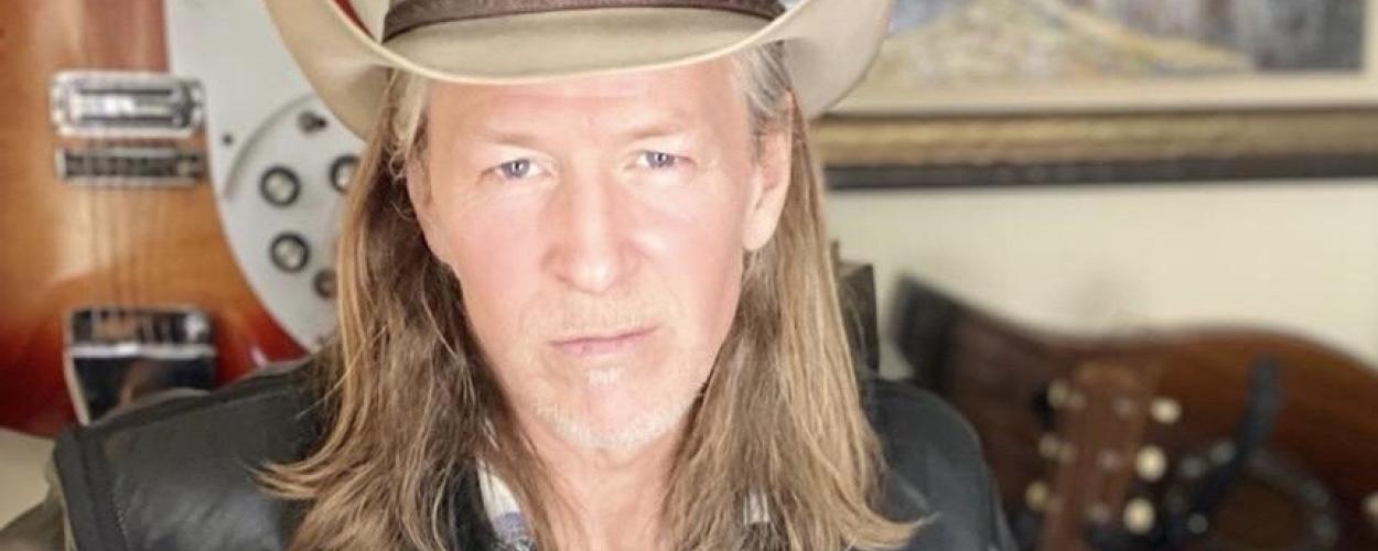 The image is a medium shot portrait of a middle-aged man with long, blonde, wavy hair, wearing a light brown cowboy hat. He has a serious expression and is looking directly at the camera. He is wearing a dark leather jacket over a blue and white plaid shirt.