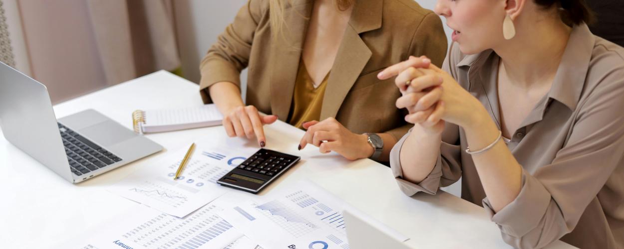 Two people sit at a desk with a laptop, paper files, and a calculator.