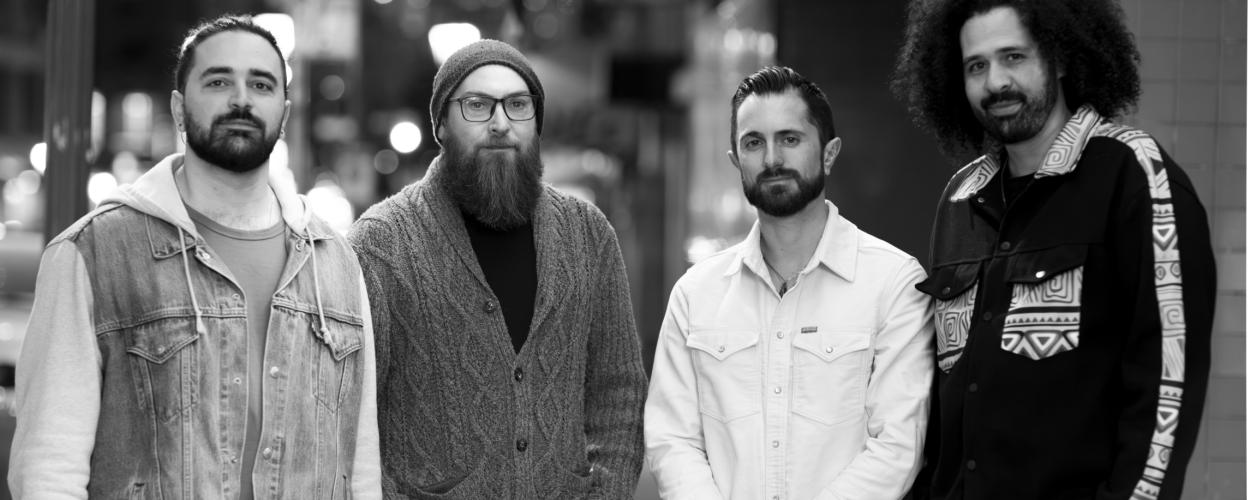 A black and white, medium shot, eye-level photograph features four men standing in what appears to be an urban environment at night. The background is blurred, suggesting a shallow depth of field, and consists of out-of-focus lights and building structures, creating a bokeh effect. The lighting appears to be ambient city light, casting soft shadows and highlighting the subjects.