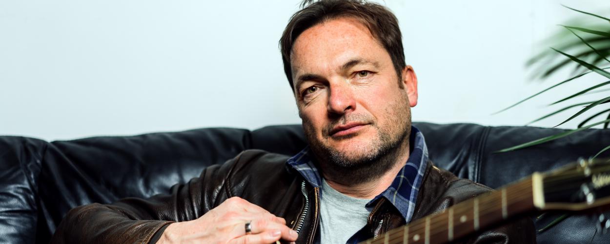 A medium shot portrait features a middle-aged man with fair skin, light brown hair, and a slight stubble. He is seated on a black leather couch, facing directly towards the camera with a neutral, contemplative expression. His eyes, a light brown color, are looking straight ahead. He has a few freckles visible on his cheeks and forehead.