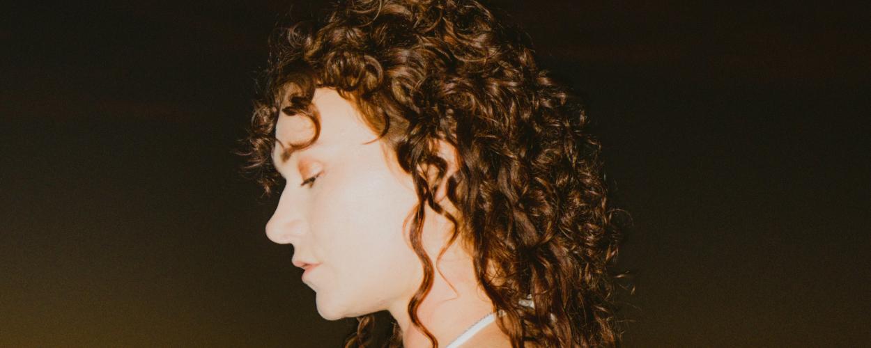 The image is a close-up, profile view of a young woman with voluminous, dark curly hair. Her head is tilted downwards, and her eyes are closed, suggesting a moment of reflection or peace. Her skin tone is fair, and she appears to be wearing subtle makeup, with a hint of eyeshadow. A delicate silver necklace is visible around her neck, and the top of a white garment, possibly a tank top or dress, can be seen.