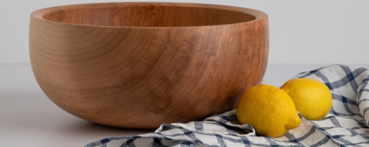 The image is a still life featuring a large, light-colored wooden bowl, two bright yellow lemons, and a blue and white checkered cloth. The setting appears to be a bright, minimally decorated studio with a white or light gray background and a white surface for the objects.