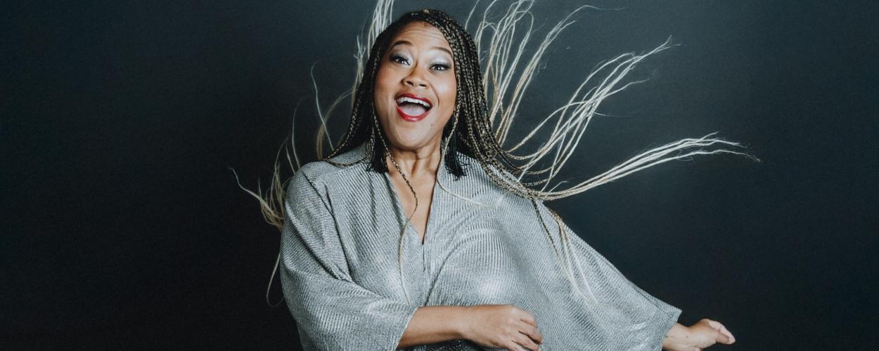 A woman is captured in a dynamic, mid-movement shot against a dark, neutral background. She appears to be of African descent and is wearing a shimmering, silver, V-neck dress with wide, flowing sleeves. Her expression is joyful and energetic, with her mouth open as if she is laughing or exclaiming. Her eyes are wide and her makeup is dramatic, with bold eyeliner and bright red lipstick.