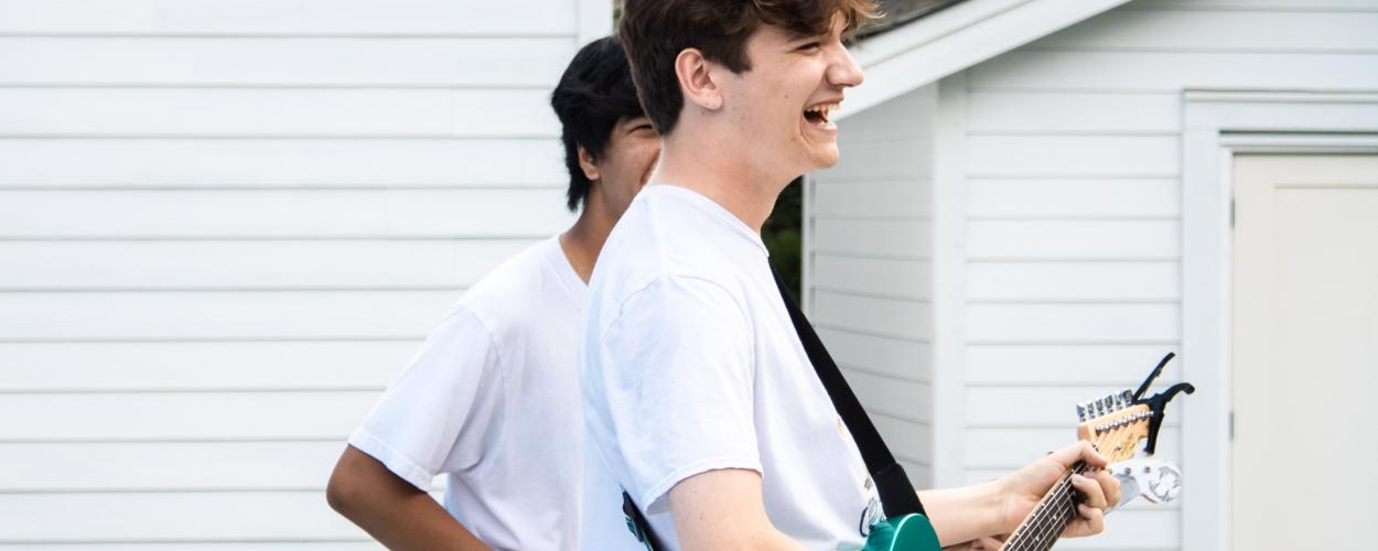 The image captures two young men outdoors, positioned in front of a white clapboard-sided building, likely a residential structure. The composition is dynamic, with the primary subject—a young man with tousled brown hair—occupying the right side of the frame. 