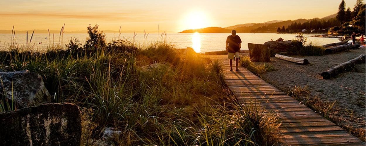 A man walks away from a waterfront along a wooden boardwalk as the sun sets behind him.