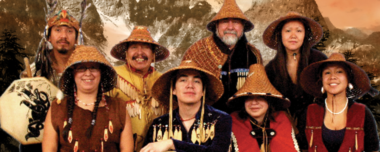 The image is a composite that features a group of nine people, likely Indigenous performers, arranged in two rows against a dramatic backdrop of snow-capped mountains and a fiery, sunset-like sky. The overall color palette is dominated by warm, earthy tones of brown, orange, and gold, with accents of dark green from the pine trees and the blue and red of the masks and some clothing.