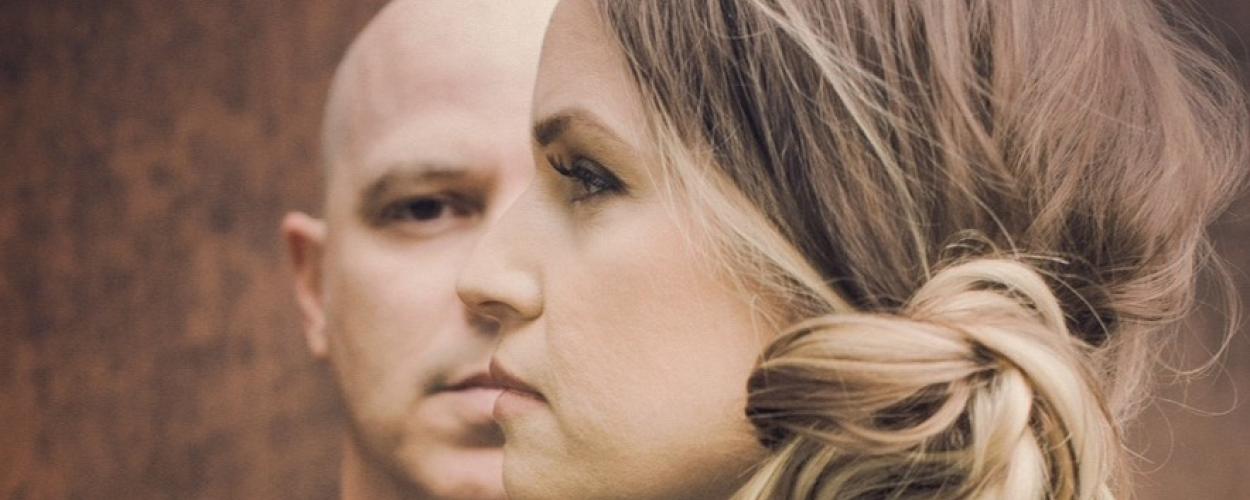 The image is a close-up, profile shot of two people, a man and a woman, superimposed or layered in a way that suggests a connection or duality. The man is bald and in the background, seen from the nose up, facing left. His expression is neutral. The woman is in the foreground, facing left, with her head angled slightly downwards. Her hair is light brown with blonde highlights, pulled back into a messy bun at the nape of her neck. Her makeup is visible, with eyeliner and mascara accentuating her eyes. 