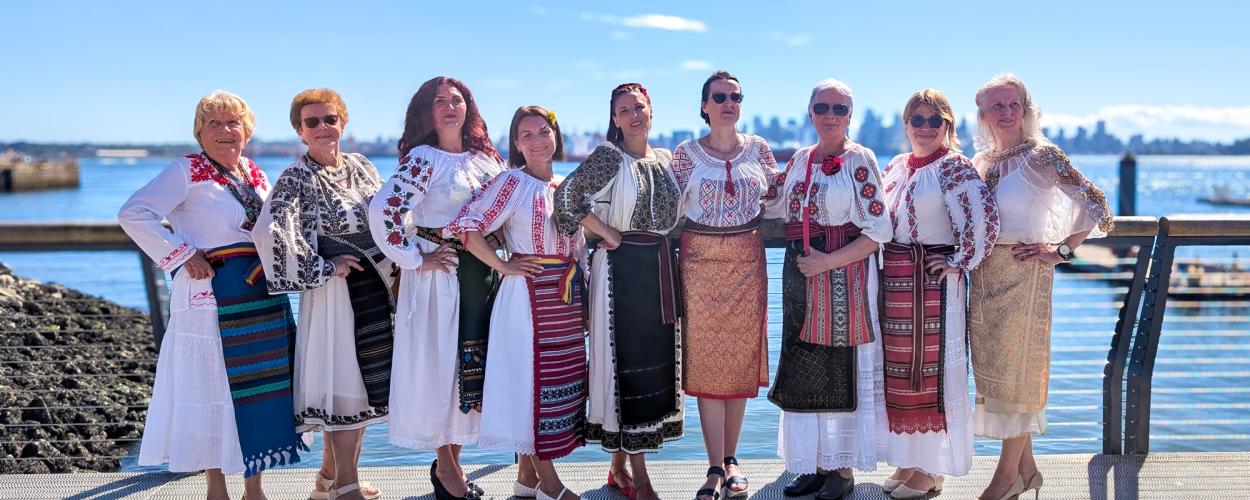 A group of nine women, all of varying ages and ethnicities, are posing for a photo outdoors on a sunny day. They are standing on a boardwalk or pier, with a body of water and a city skyline in the background. The women are dressed in traditional, elaborately embroidered folk costumes. The costumes are predominantly white with intricate red, black, and blue embroidery on the sleeves, necklines, and hems. Many of the women are wearing layered skirts and sashes, also adorned with traditional patterns and brigh