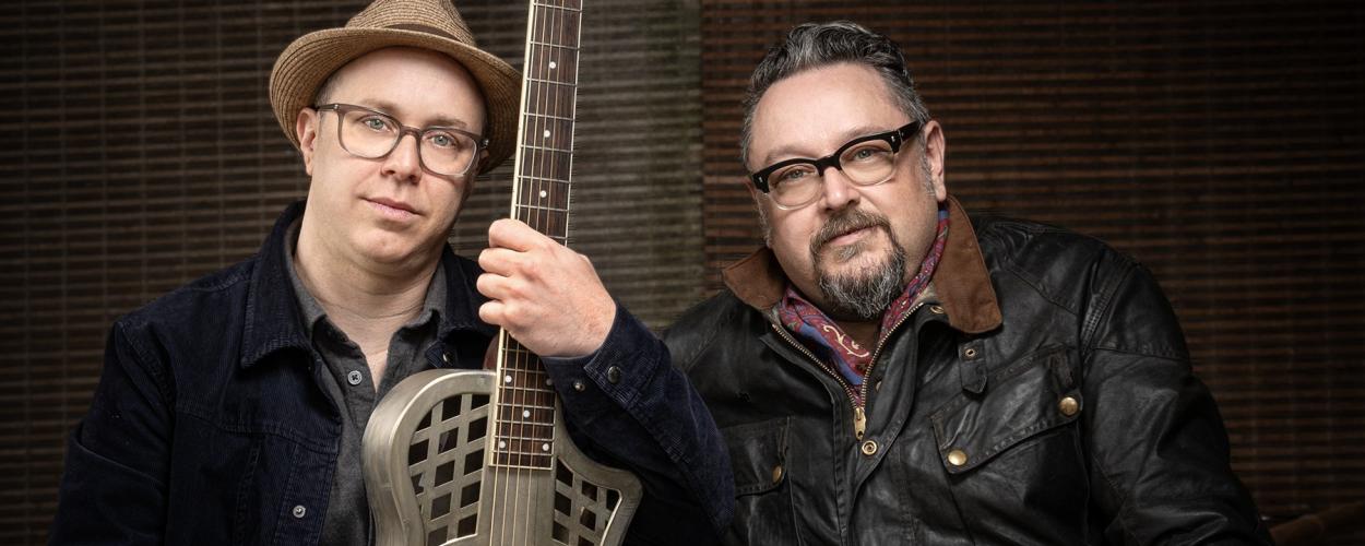 The image is a medium shot portrait of two middle-aged men, positioned side-by-side and looking towards the camera. They are both wearing glasses and appear to be musicians, as one of them is holding a resonator guitar.