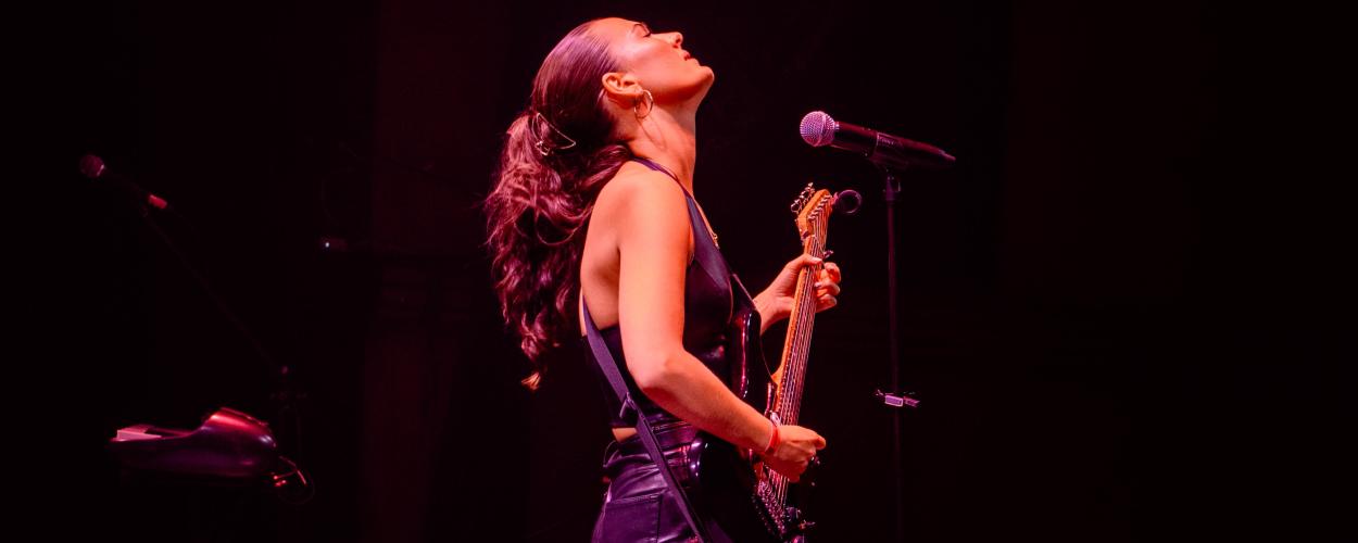 The image captures a dynamic moment of a female musician performing on stage. She is positioned in the center-left of the frame, looking upwards with her head tilted back, her eyes closed, exuding a sense of passionate immersion in her performance. Her dark, wavy hair is tied back into a ponytail and flows loosely around her shoulders and back.