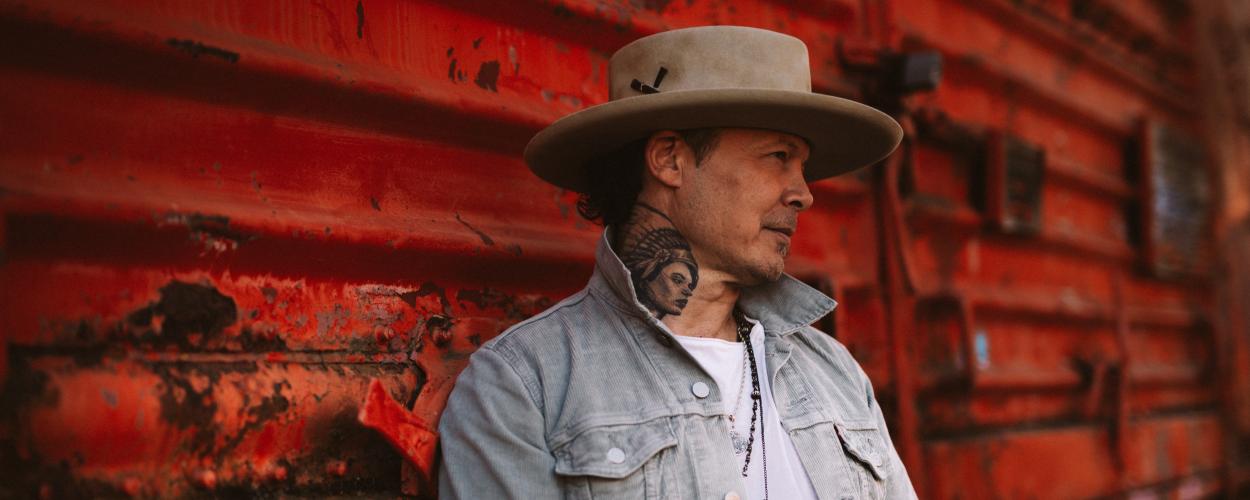 The image is a medium shot portrait of a man leaning against a weathered, corrugated red surface. The man is wearing a light grey, possibly corduroy, jacket over a white t-shirt. He has on a wide-brimmed, light brown hat. A prominent tattoo of a Native American headdress and profile is visible on his neck, extending upwards towards his ear. He is also wearing a necklace with multiple dark beads and a turquoise pendant.