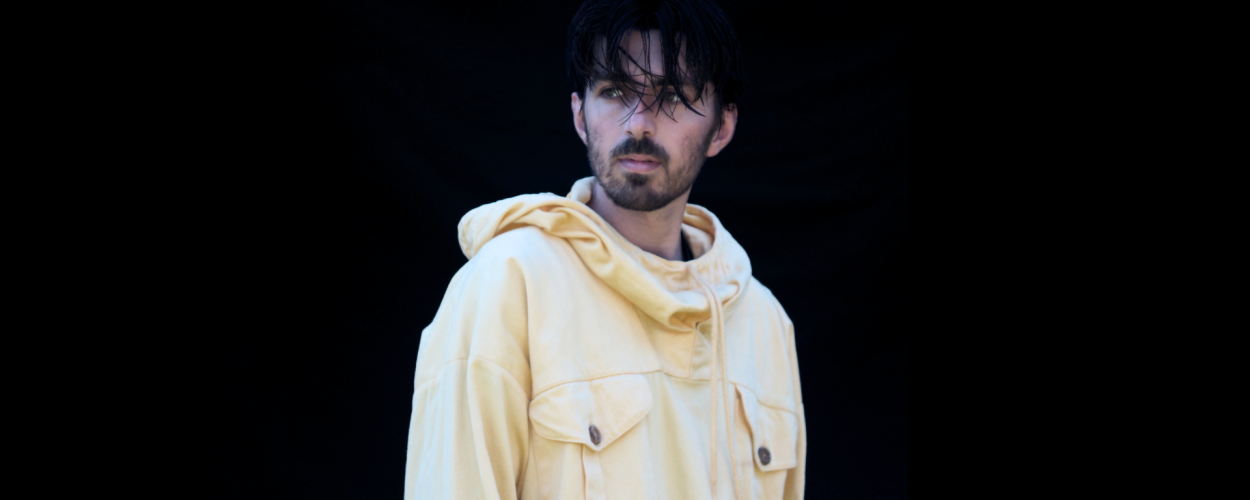 A man with dark, somewhat messy hair and a beard is shown from the chest up, looking to his left with a neutral expression. He is wearing a light yellow, oversized hooded jacket with a drawstring and two large buttoned pockets on the front. The jacket appears to be made of a sturdy material like canvas or denim. The background is completely black and featureless, creating a stark contrast with the man and his clothing. The lighting highlights the textures of the fabric and the contours of the man's face and