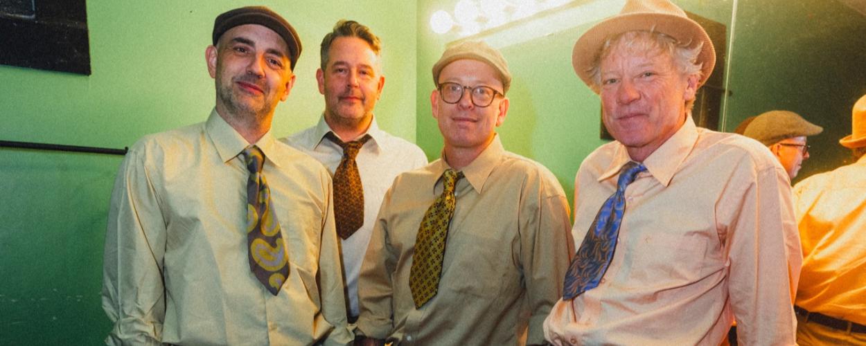 The image features a group of four middle-aged men, dressed in retro-style attire, posing for a photograph. They are all wearing light-colored button-down shirts and ties, with variations in their hats and pants. The man on the far left wears a flat cap and a tan shirt with a colorful tie featuring a paisley-like pattern in yellow, grey, and brown. He also sports light grey pleated pants and a dark belt, with his left hand in his pocket. The second man from the left wears a dark patterned tie and a white sh