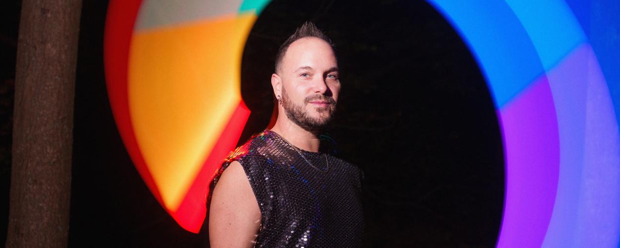 The image is a medium close-up portrait of a man standing in front of a blurred, circular light display at night. The man is looking directly at the camera with a slight smile. He has short, spiky dark hair, a beard, and is wearing a sleeveless, black sequined top and a silver chain necklace. His left arm is slightly extended, and he appears to be holding something small and dark in his hand.
