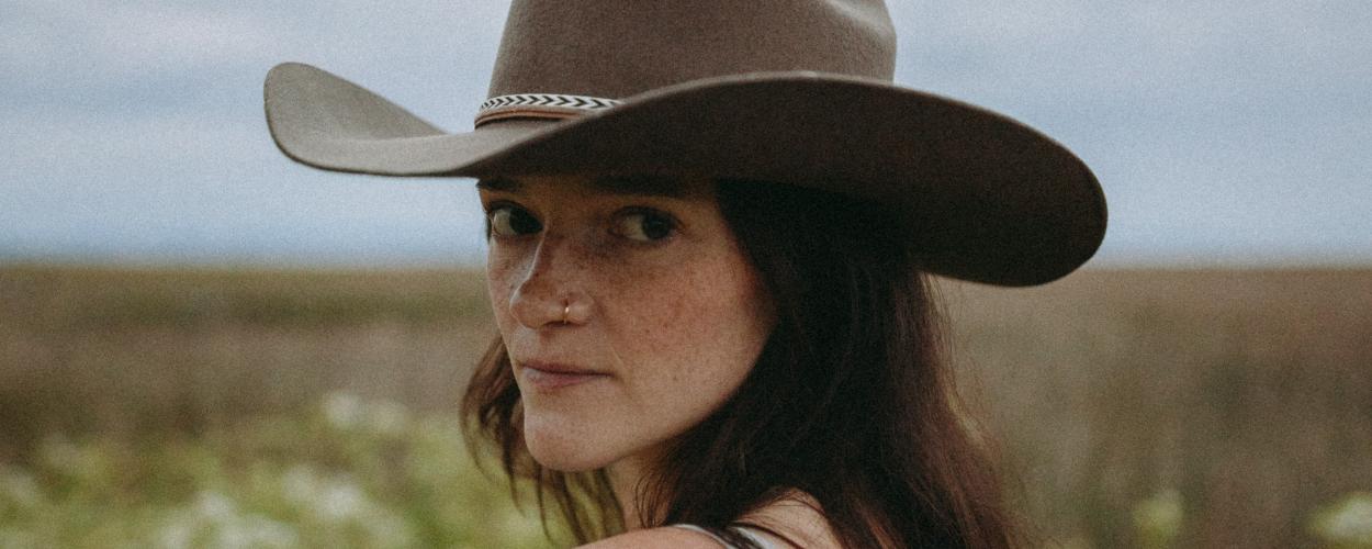 The image is a close-up portrait of a young woman wearing a wide-brimmed, dark brown cowboy hat. She is looking over her left shoulder towards the camera, with her face angled slightly to the right. Her expression is calm and direct, with a hint of a gaze.