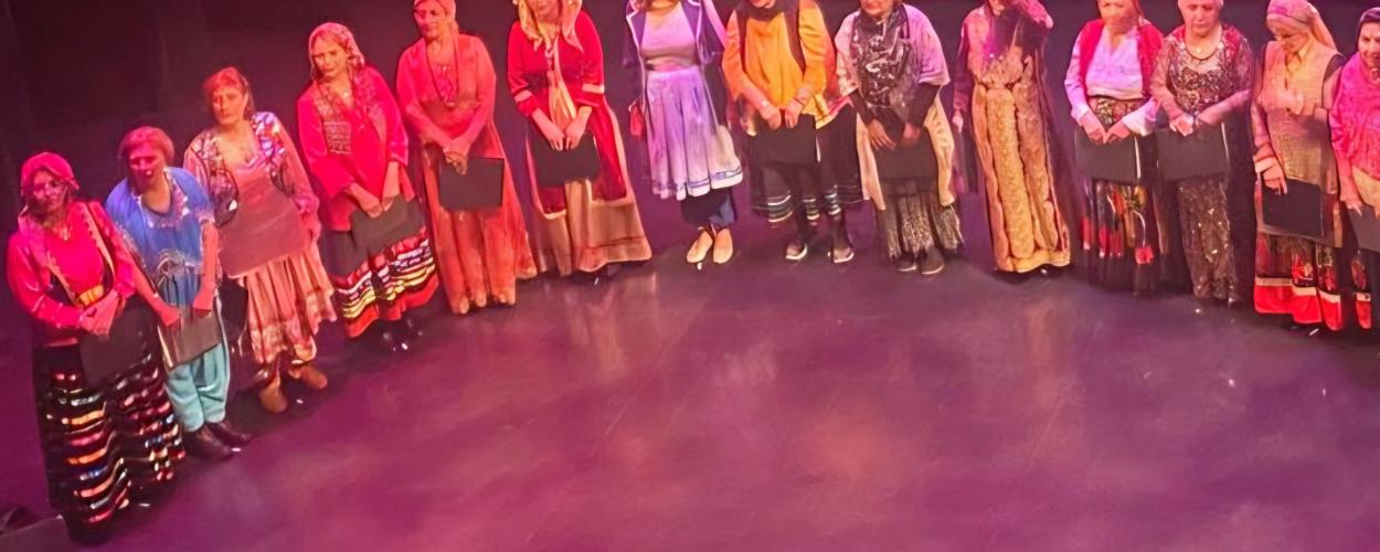 The image shows a group of twelve women standing in a line on a stage, likely after a performance or ceremony. They are all dressed in traditional, colorful clothing, and most of them are holding black folders or certificates. The women are positioned from left to right across the frame, forming a diagonal line that recedes into the background. The stage floor is a deep, rich magenta color with subtle highlights, suggesting stage lighting. The background is dark and out of focus, with a faint, abstract patt