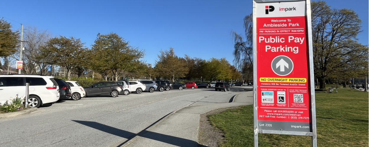 A red parking sign reads "Welcome to Ambleside Park - Public Pay Parking" with a parking lot and blue sky in the background.