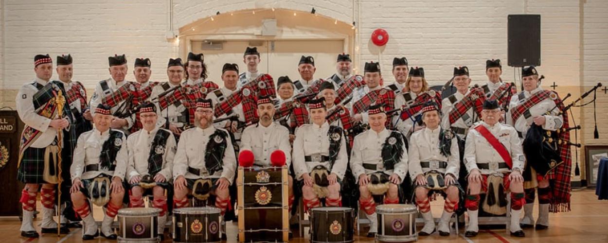 A pipe band poses for a group photo in a gym, showcasing their uniforms and instruments with smiles and camaraderie.