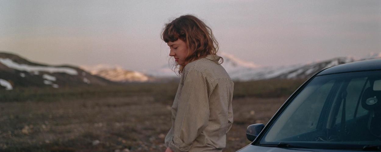 A young woman stands in profile beside the front of a dark-colored car, positioned slightly to the right of the frame. She has shoulder-length, wavy brown hair with bangs, and her head is tilted downward, her gaze directed toward the ground, suggesting introspection or melancholy.