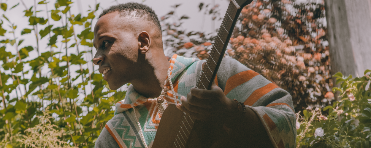 The image features a close-up, profile view of a Black man holding a guitar. He is wearing a poncho with a geometric pattern in shades of teal, orange, and grey. His expression is one of concentration or perhaps enjoyment, with his mouth slightly open and a smile visible. His skin tone is dark. He has short, textured hair.