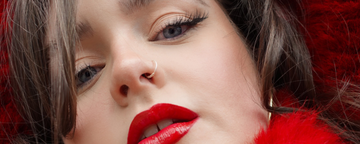 The image is a close-up, low-angle portrait of a young woman's face, with her head tilted slightly upwards and to the left. The foreground is dominated by her face, with the background largely obscured by vibrant, fluffy red material, possibly faux fur trim or a scarf.