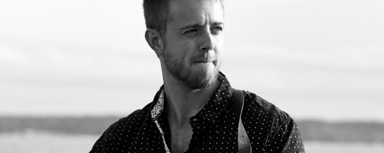 This is a medium shot, black and white photograph of a man holding a guitar. He is positioned slightly off-center to the left, with his head turned to the right, looking away from the camera. The man has light-colored hair styled upwards and a short, well-maintained beard. He is wearing a dark shirt with a repeating pattern of small white triangles or dots. The collar of the shirt is visible and has a contrasting white, patterned fabric. A dark leather strap from the guitar is visible across his chest.